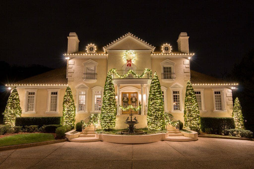 A large home with its rooflines, gables and front trees outlined in warm white Christmas lights at night